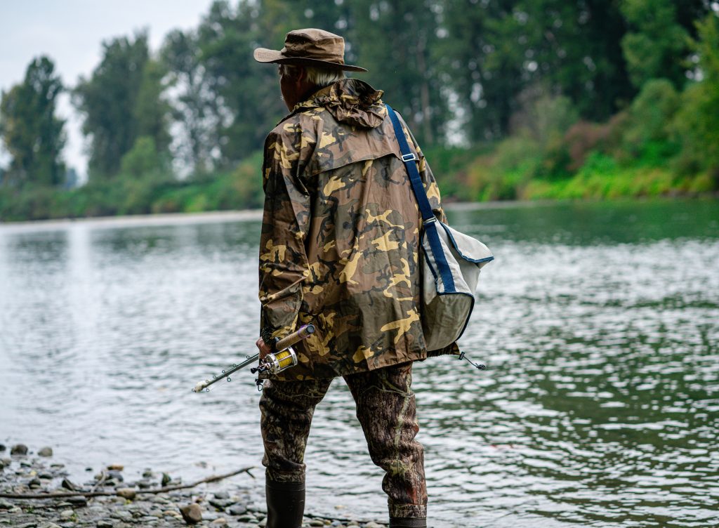 Man in camouflage top and pants walking on shore with fishing rod